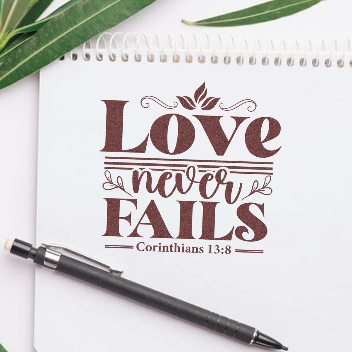 Brown custom stamp seal reading Love never fails Corinthians 13:8 on white notebook, with green leaves and pen.