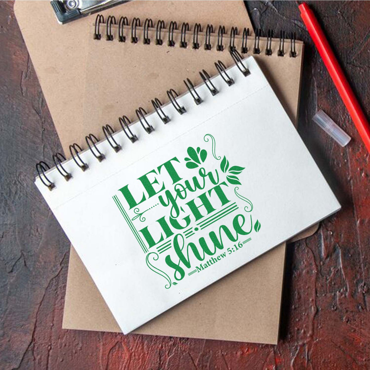 White spiral notebook with green Let your light shine scripture stamp, beside a brown notebook and red pencil.