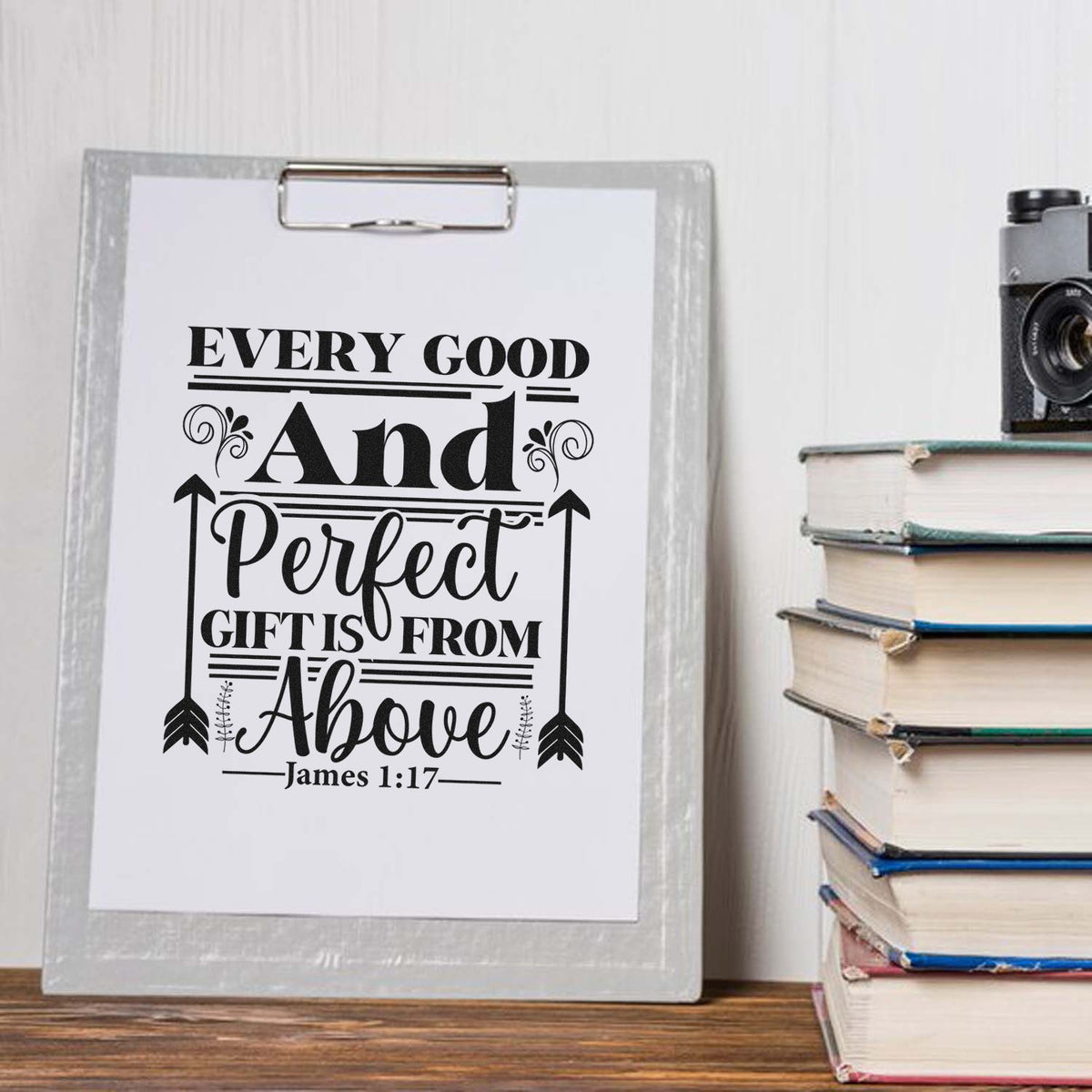 Black scripture stamp and seal design reading Every good and perfect gift is from above on a white clipboard beside stacked books.