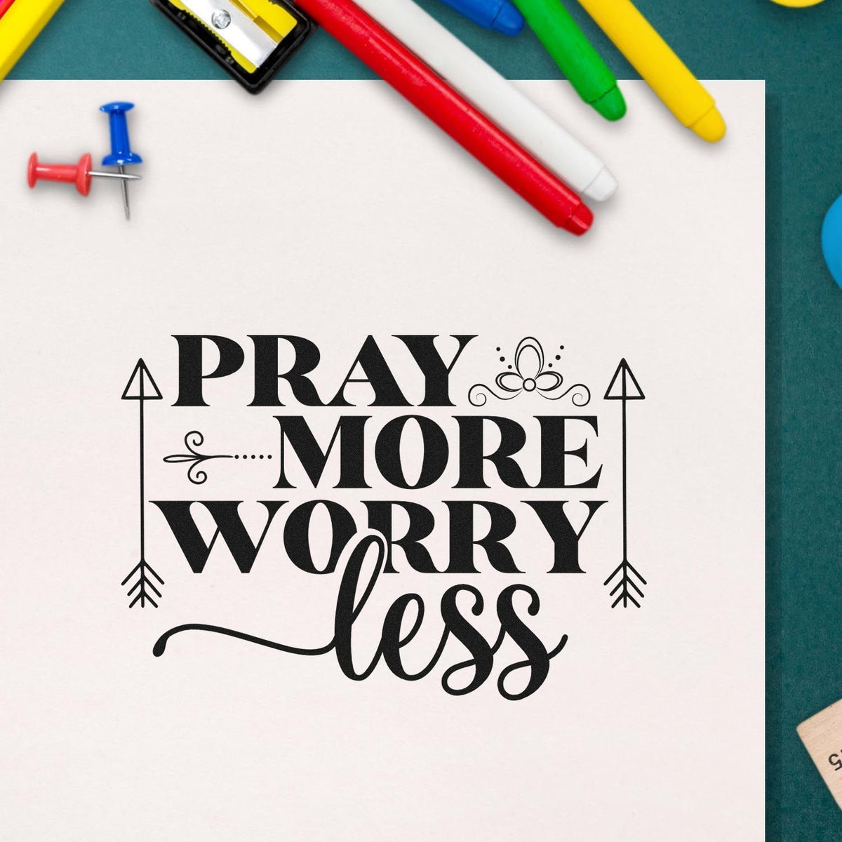 Decorative black stamp and seal reading Pray more worry less on white paper, with colorful pens and push pins.