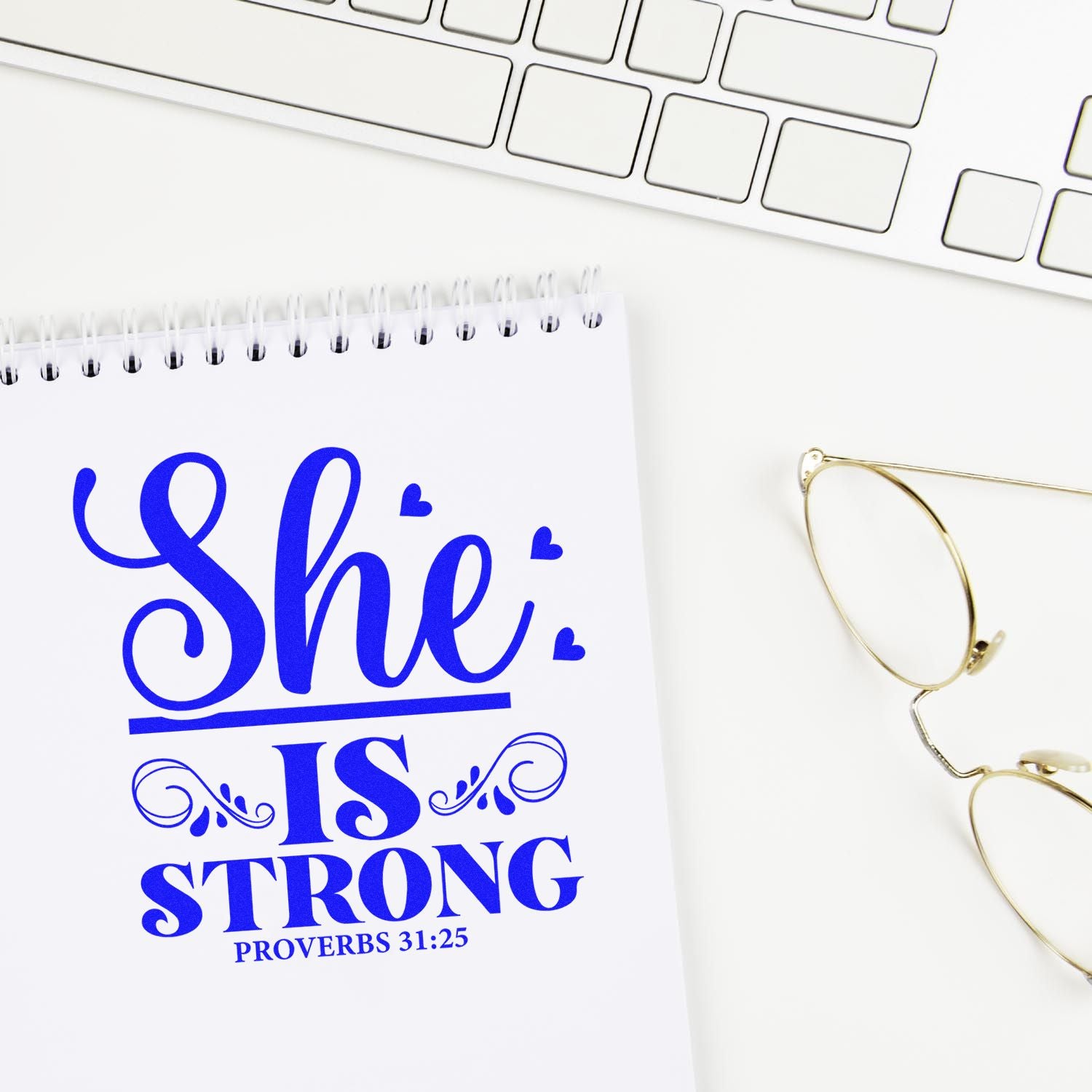 Blue She is Strong Proverbs 31:25 stamp/seal design on a white spiral notebook beside gold glasses and a white keyboard