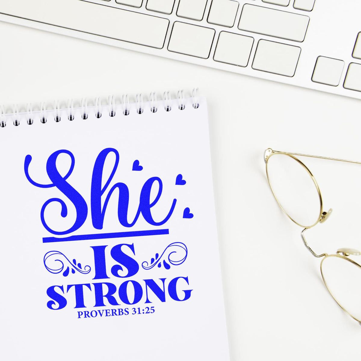 Blue She Is Strong Proverbs 31:25 stamp and seal design on white notebook beside gold glasses and keyboard.