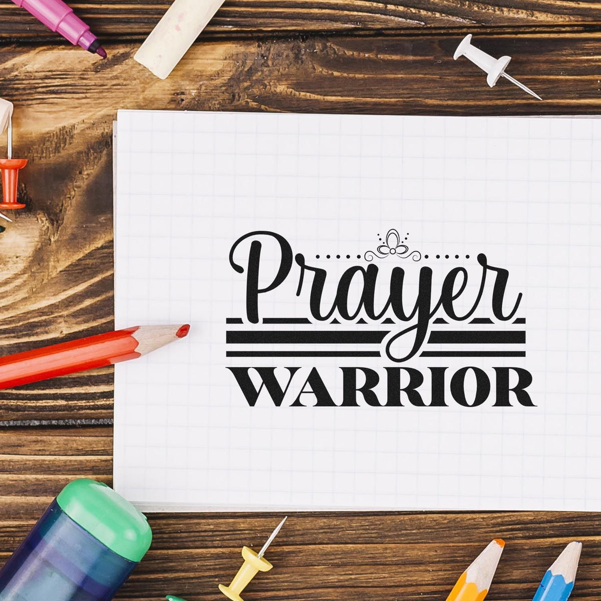 Black Prayer Warrior stamp/seal design on white grid paper, surrounded by colorful pencils and push pins on rustic wood.