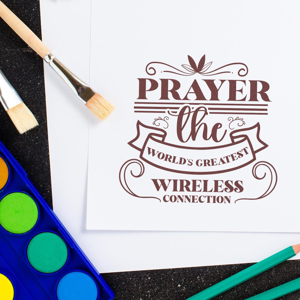 Brown decorative rubber stamp seal reading 'Prayer the World's Greatest Wireless Connection' on white paper, with brushes and paint pans.