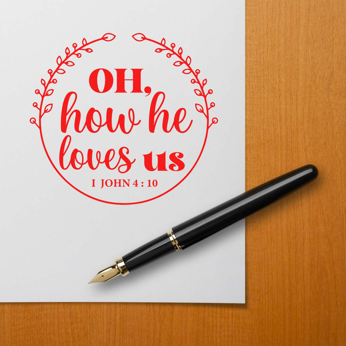 Red circular stamp seal with floral border reading OH, how he loves us and I John 4:10 beside a black fountain pen.