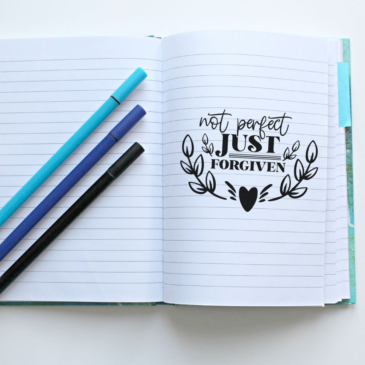 Open lined notebook with black Not perfect, just forgiven stamp seal and turquoise, purple, and black markers.