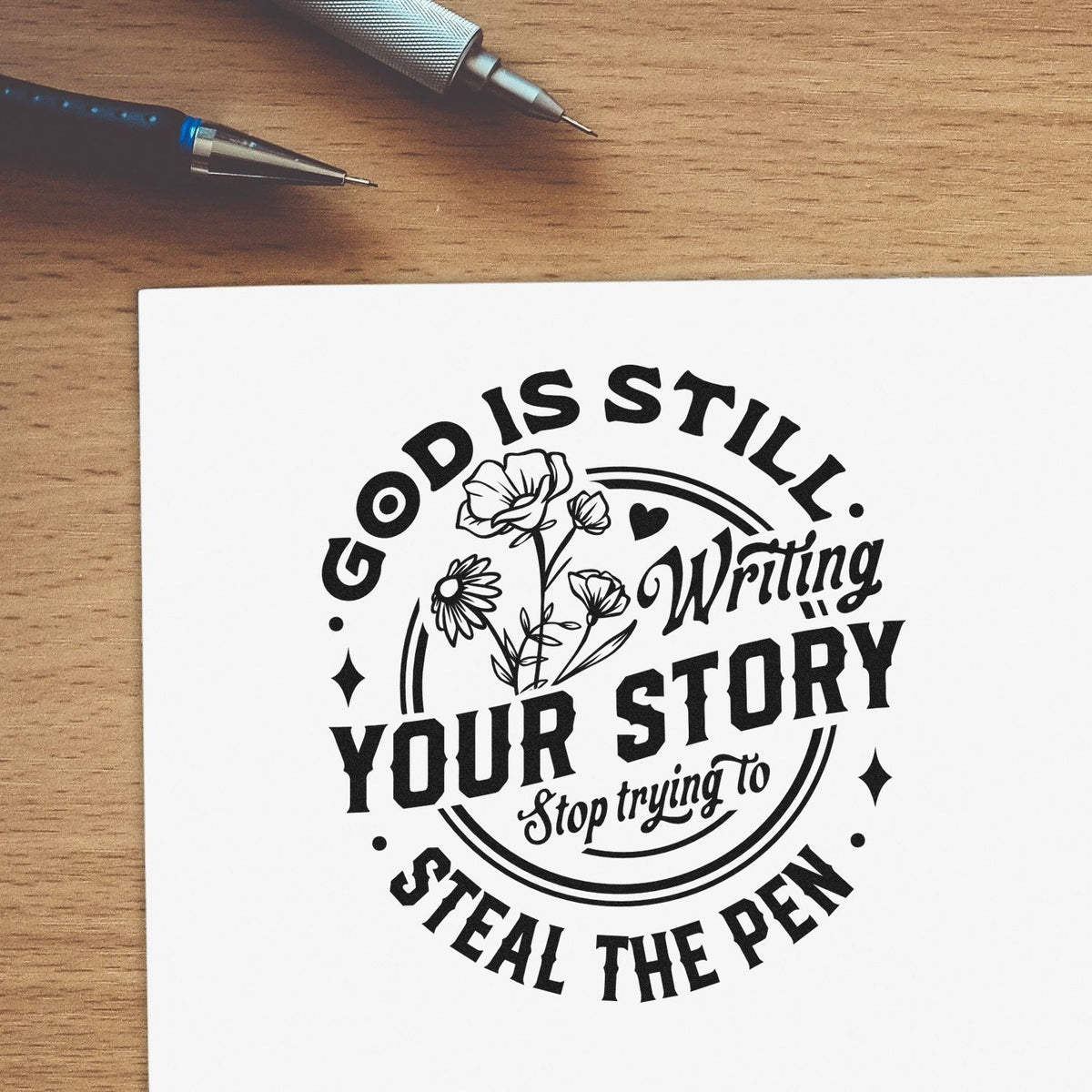 Black decorative stamp seal on white paper with floral quote, beside pens on a wood desk.