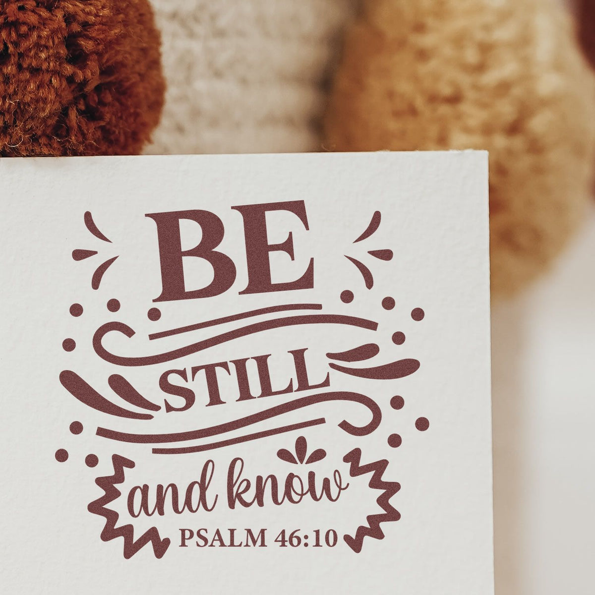 Maroon decorative stamp and seal design reading Be still and know with swirls and Psalm 46:10 on white paper.