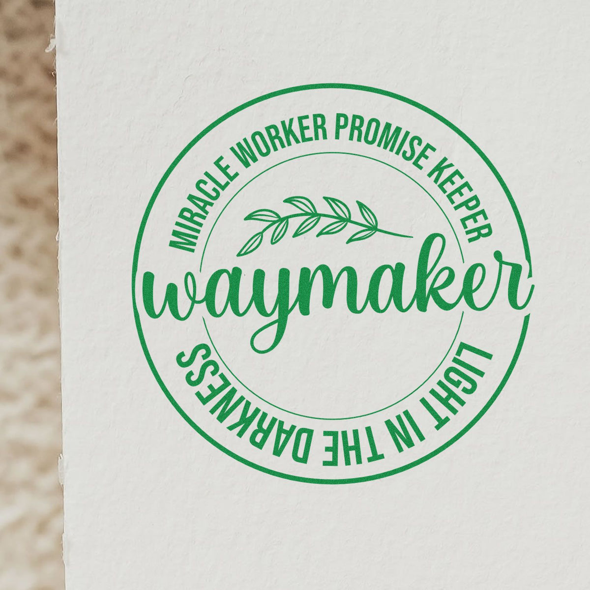 Green circular rubber stamp seal on white paper, labeled Waymaker, Miracle Worker, Promise Keeper, light in the darkness with leafy branch.