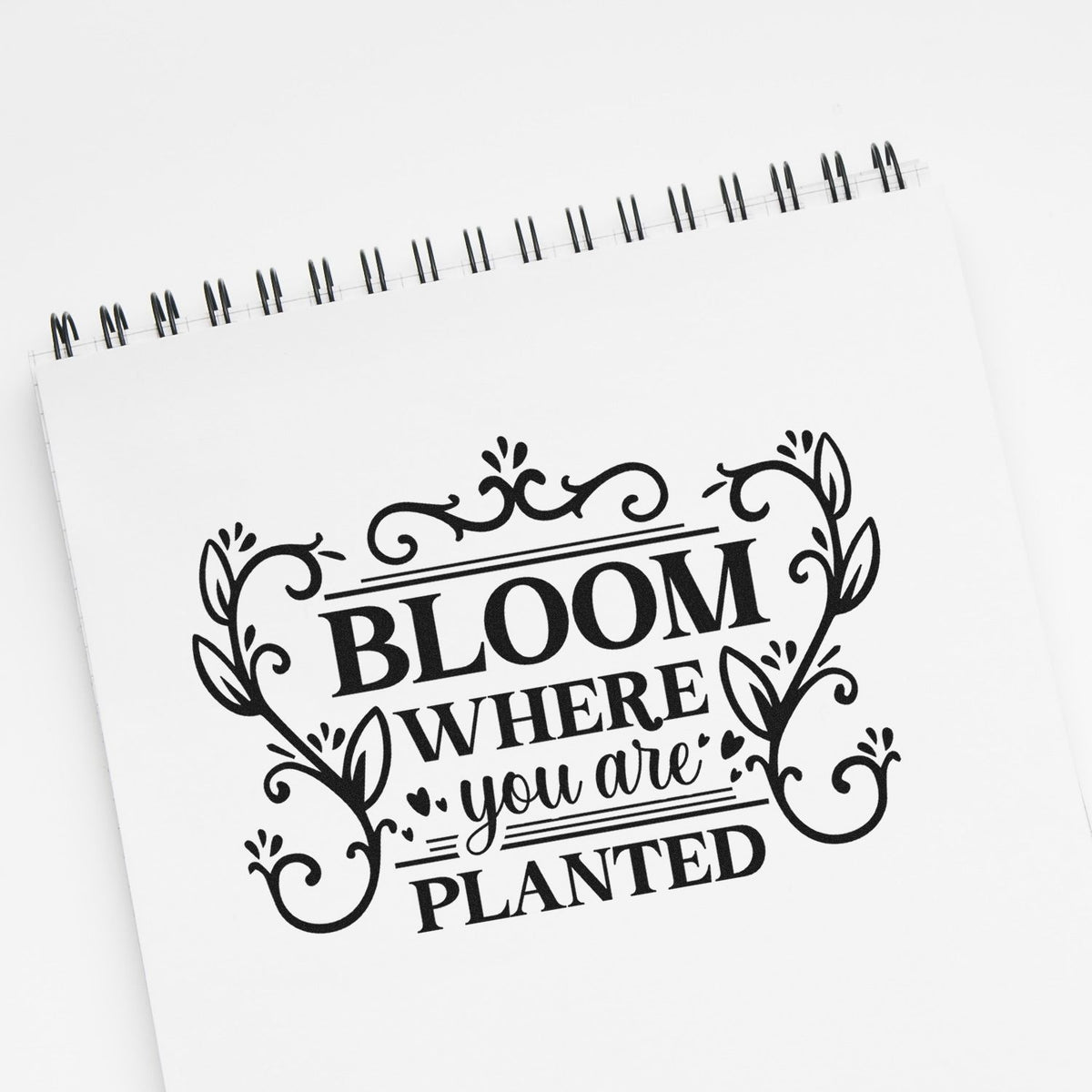 Black floral stamp and seal design reading Bloom Where You Are Planted on a white spiral notebook.