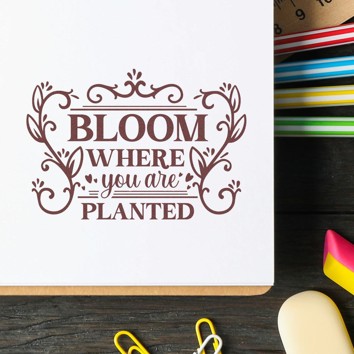 Brown decorative rubber stamp seal with floral frame and the words Bloom where you are planted on white paper.