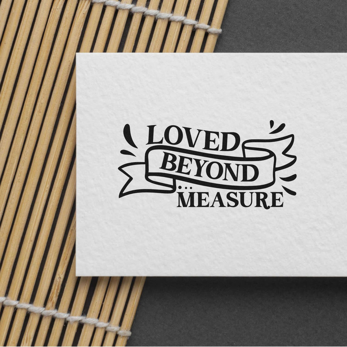 Black rubber stamp seal reading Loved Beyond Measure on white paper, set on a bamboo mat over a dark background.