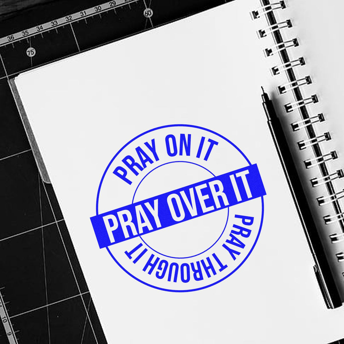 Blue stamp and seal design reading Pray on It, Pray Over It, Pray Through It on a white spiral notebook with pen.