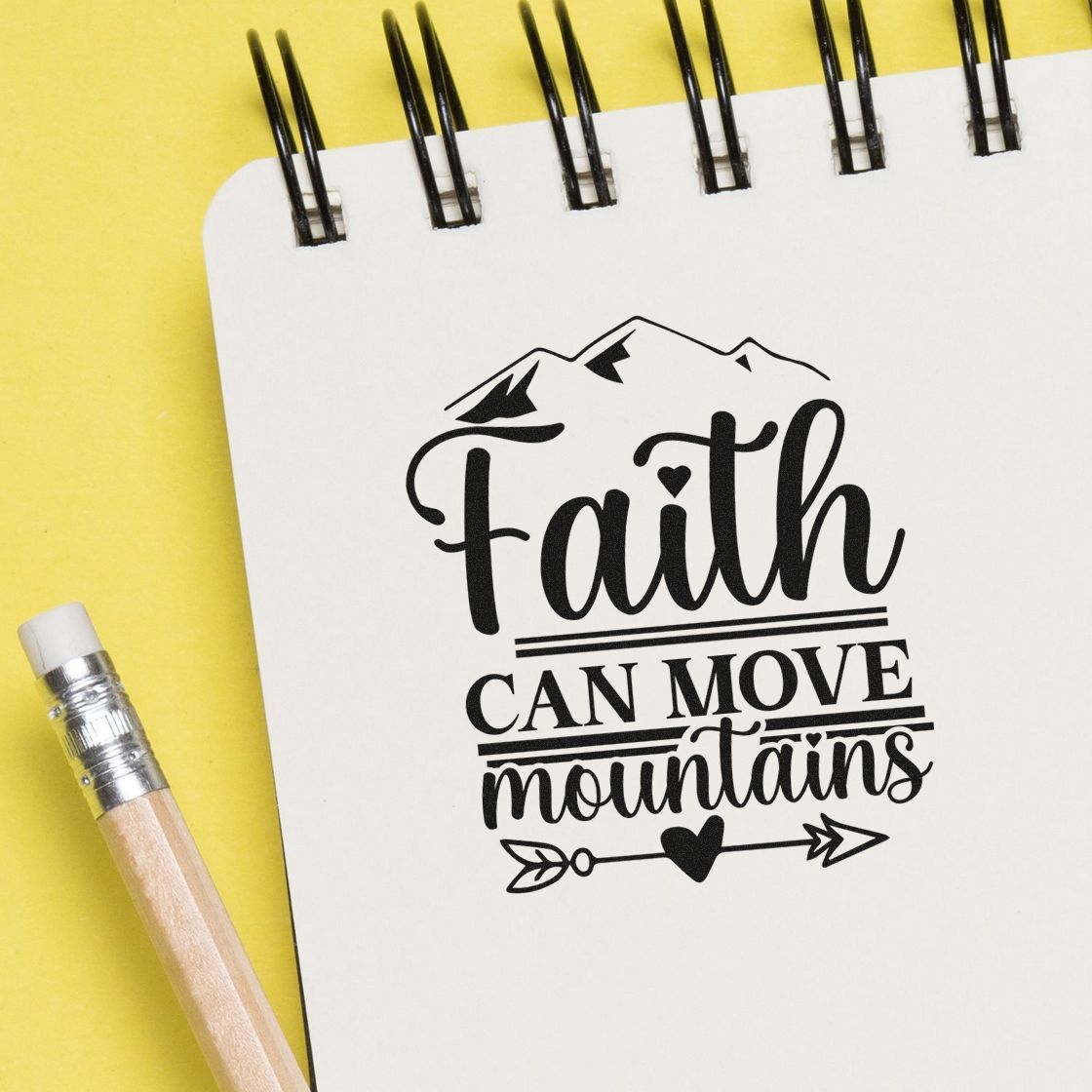 Black faith-can-move-mountains stamp and seal design on a white spiral notebook, with mountain graphic, on yellow background.