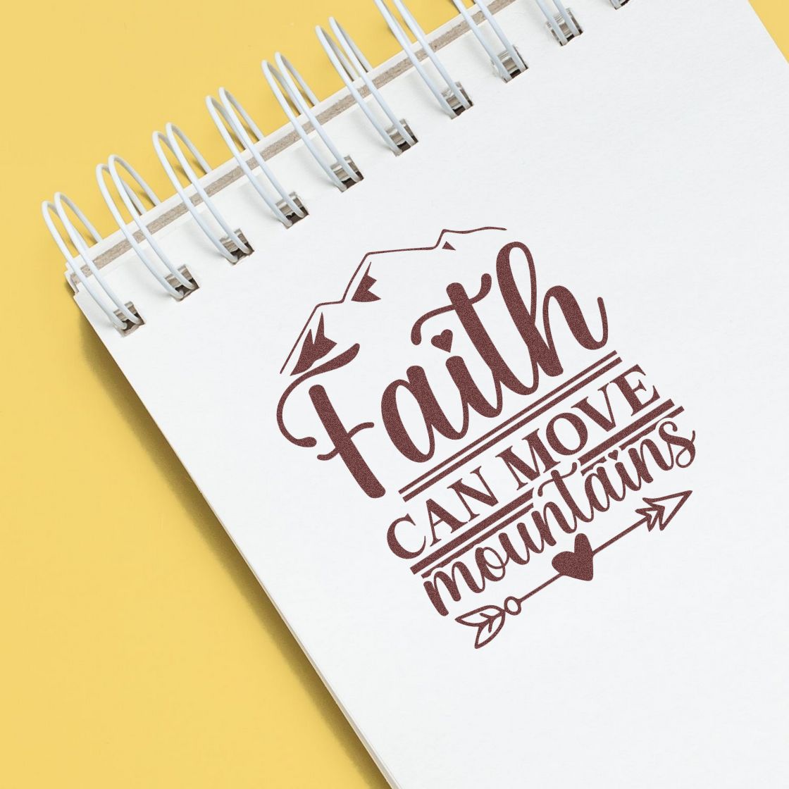 Maroon faith can move mountains stamp seal on a white spiral notebook against a yellow background.