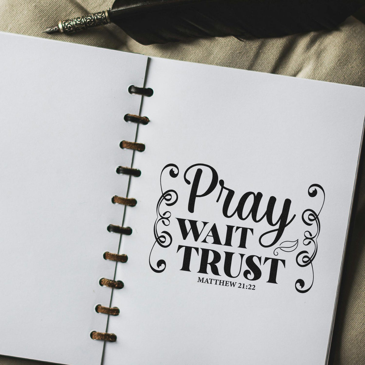 Black decorative stamp/seal reading Pray Wait Trust in an open spiral-bound notebook, with Matthew 21:22.