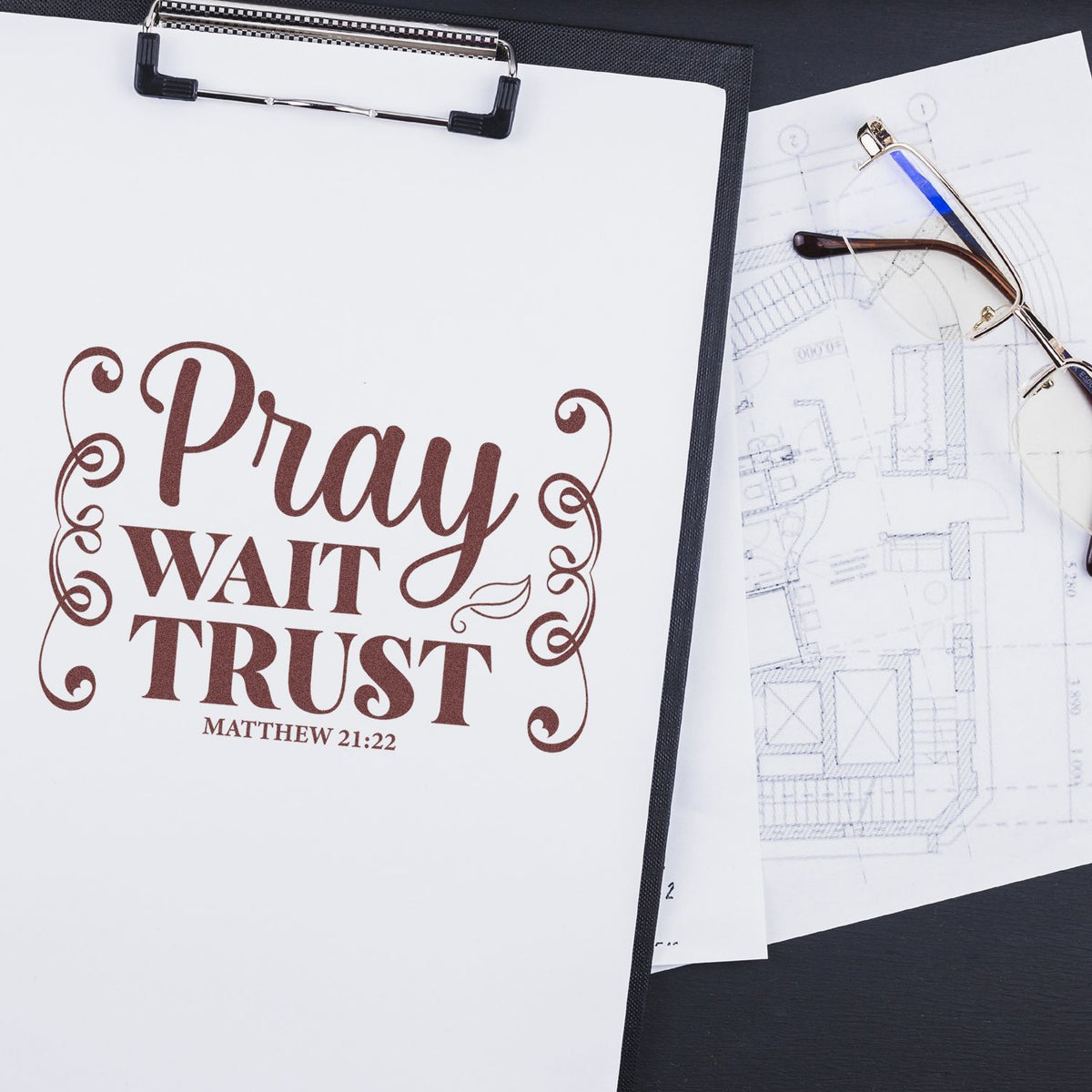 Brown decorative faith stamp/seal on white clipboard, with eyeglasses and blueprint sketches on black background.