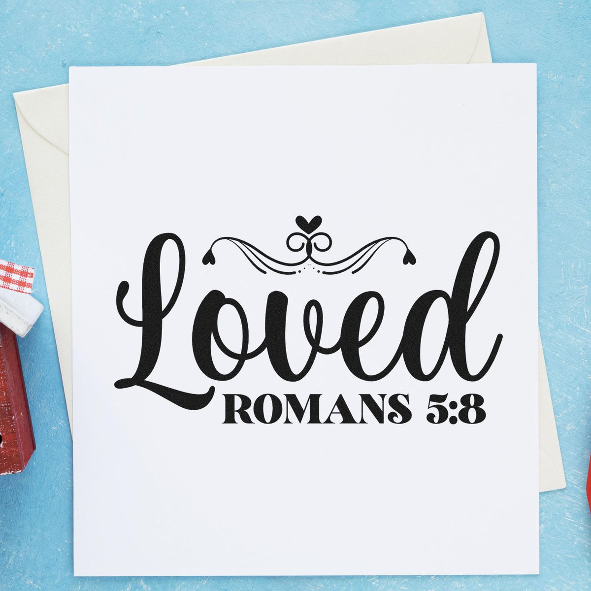 Black calligraphy Loved Romans 5:8 stamp seal on white card with decorative flourish on a blue background