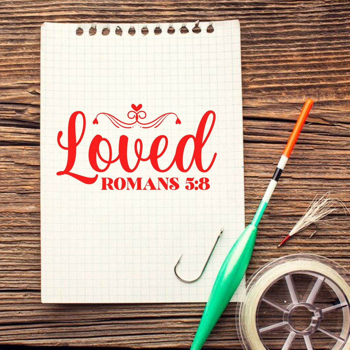 Red Loved Romans 5:8 stamp on white grid notepad with green tool, wood table, and tape roll seal products.
