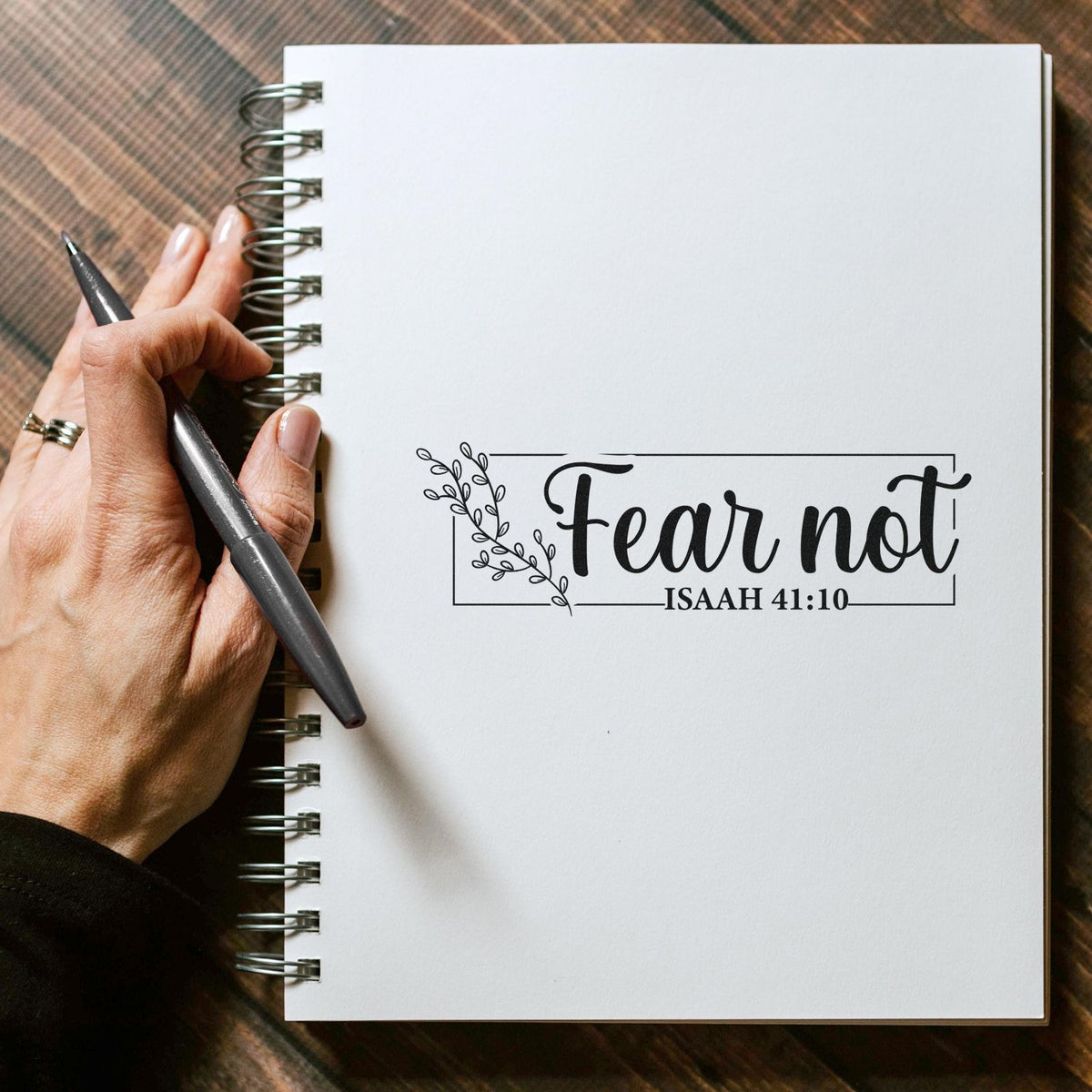Black decorative stamp/seal reading Fear not Isaiah 41:10 on a white spiral notebook, with a hand holding a pen.