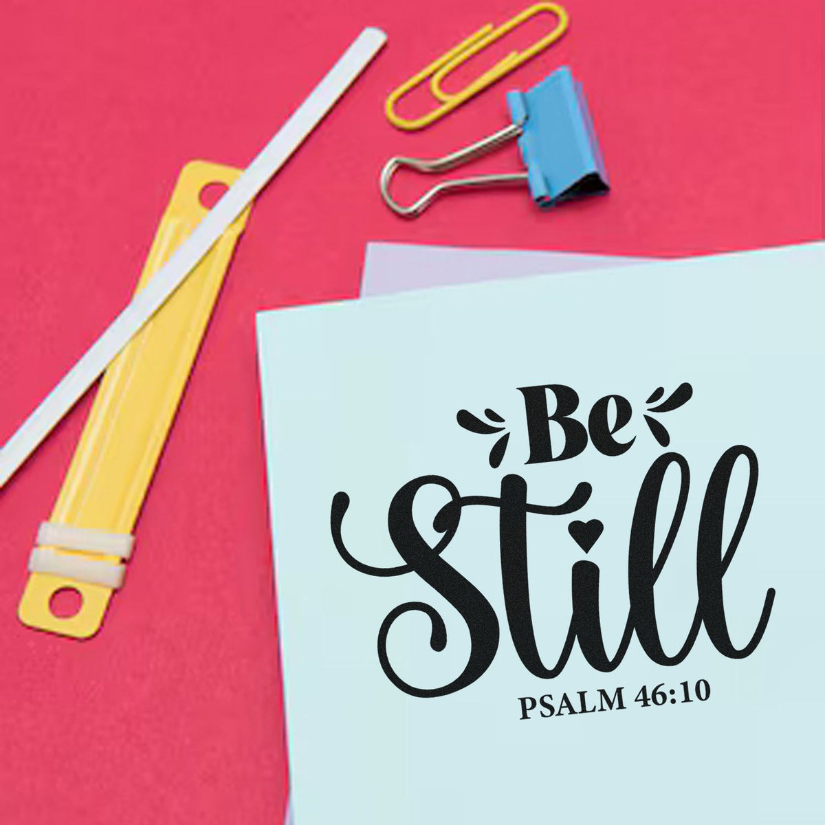 Religious stamp and seal product on pastel blue card with black Be Still verse, pink background, and colorful clips.