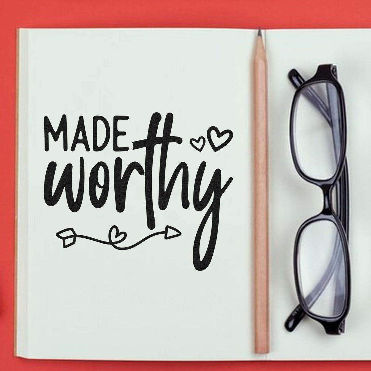 Black Made worthy stamp and seal design with hearts on a cream notebook, beside a pencil and black glasses on a red background.