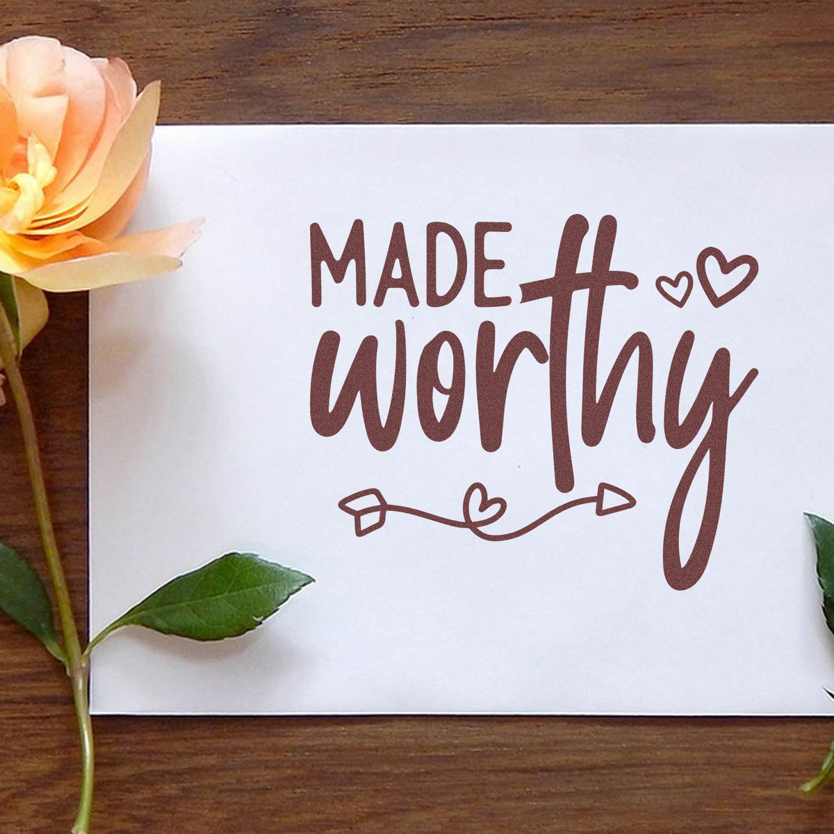 Brown Made worthy stamp seal on white paper with hearts, beside peach flower and green leaves on wood.