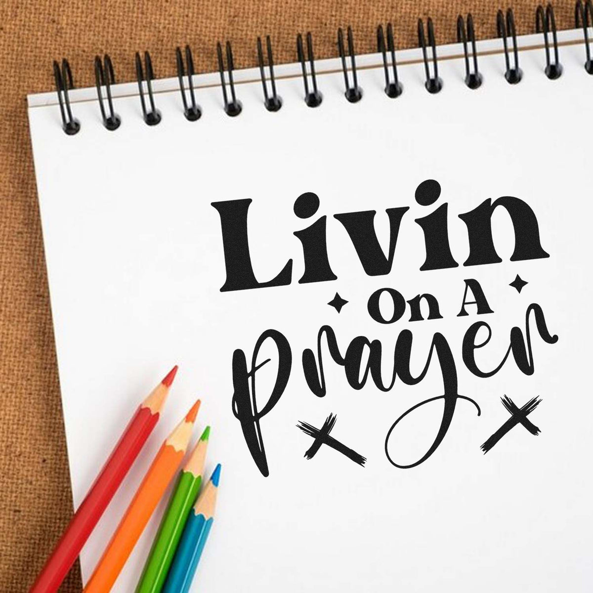 Black custom stamp and seal design on white spiral notebook reading Livin on a Prayer, with colorful pencils on a brown background.