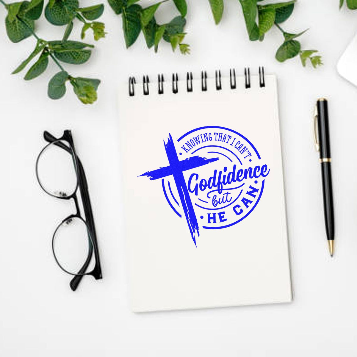 Blue Godfidence faith seal stamp with cross on white spiral notebook, beside black glasses, pen, and green leaves.