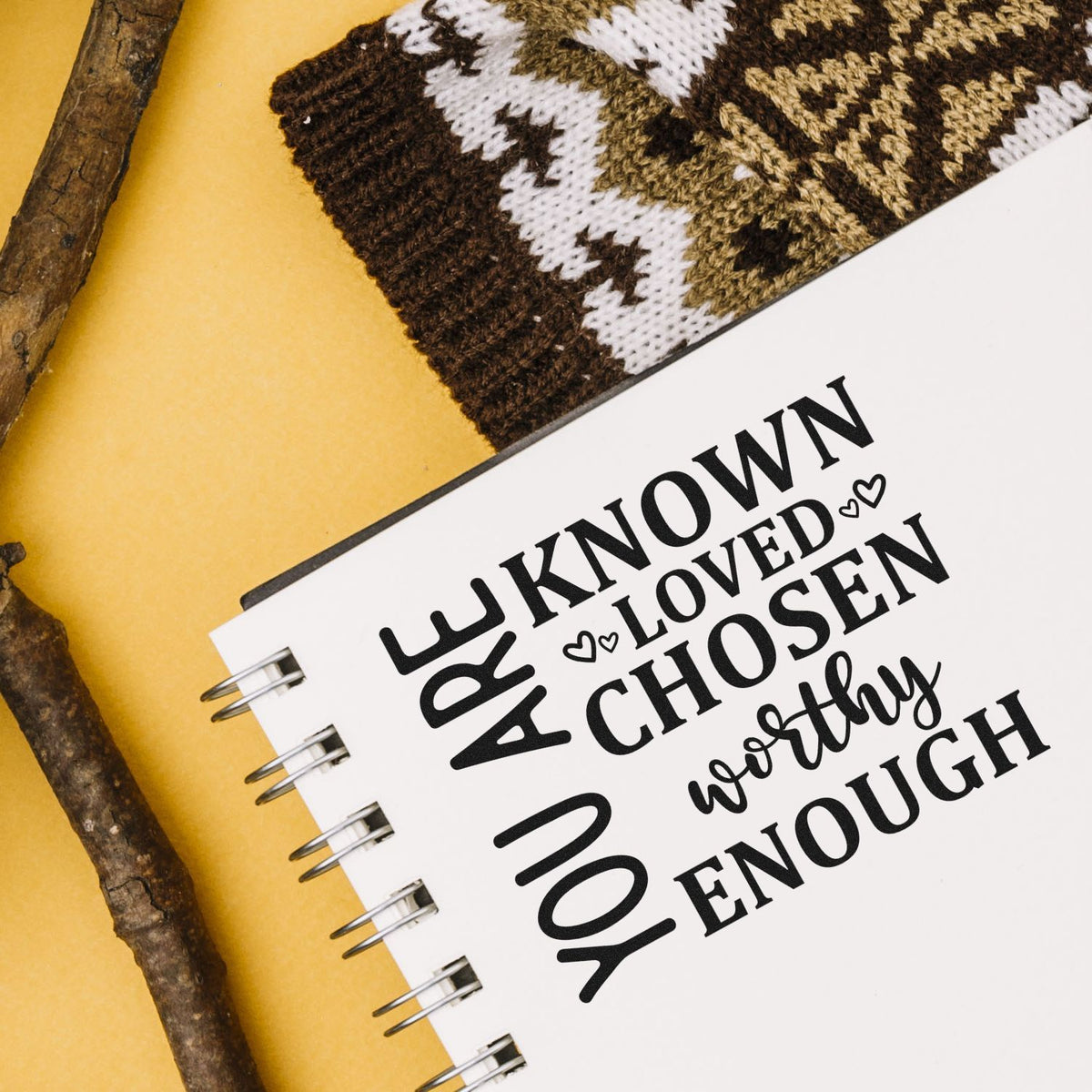Stamp and seal products with bold black inspirational words on a white spiral notebook, yellow background, and knit swatch.