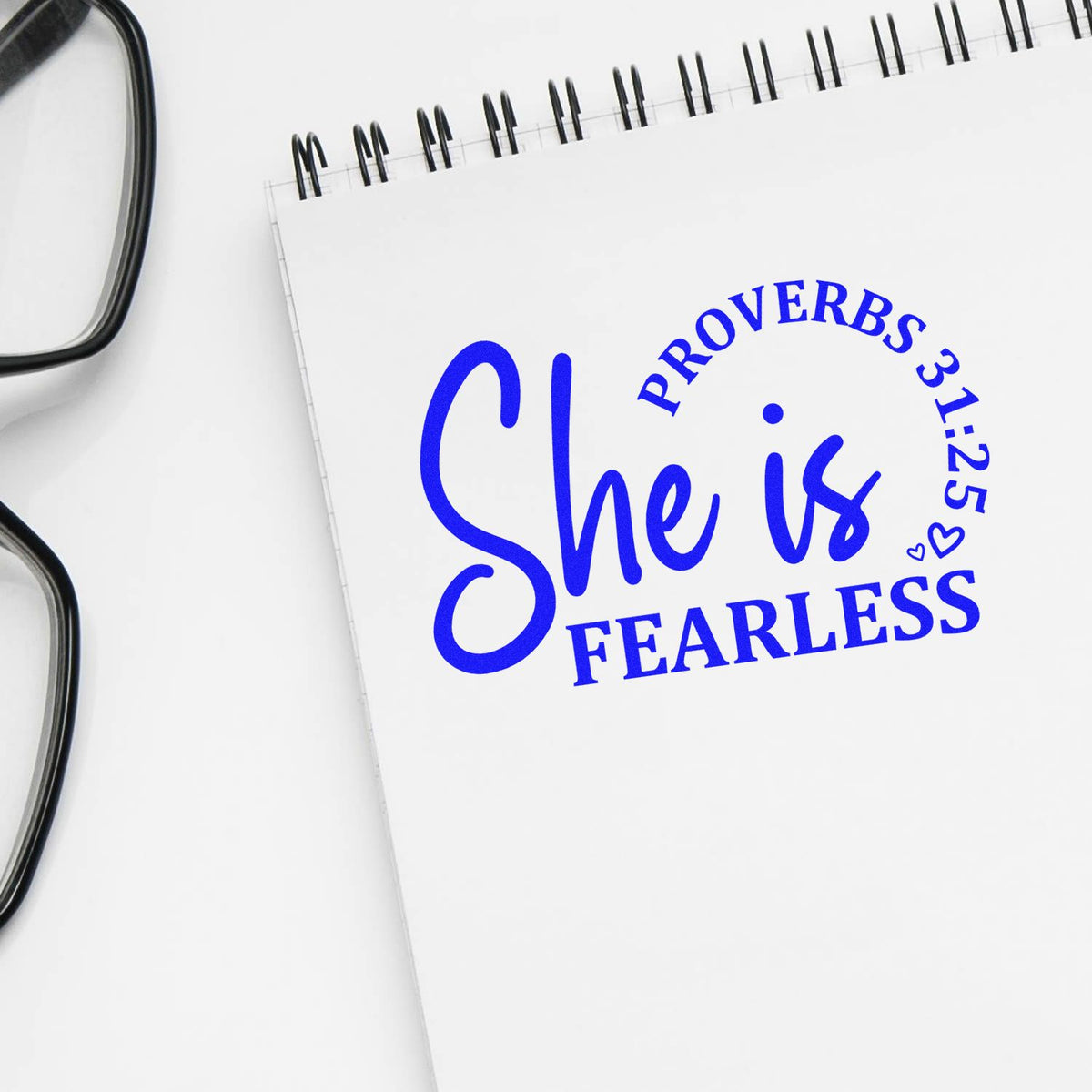 Blue stamp and seal on white spiral notebook: She is fearless with Proverbs 31:25, beside eyeglasses.