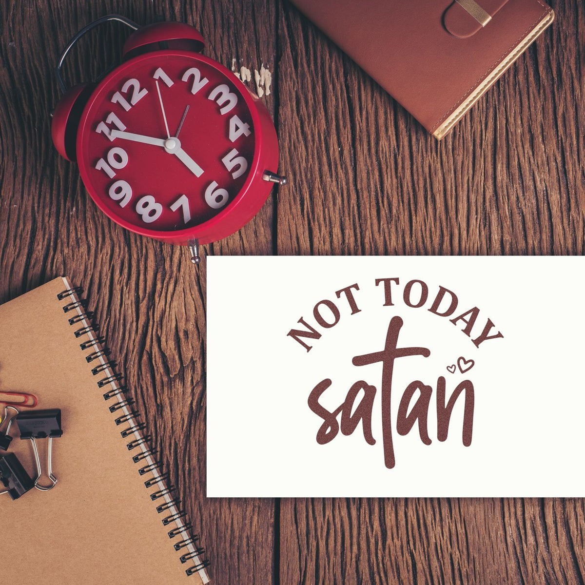 Red alarm clock, brown notebooks, and white Not Today Satan stamp and seal on a wood table.