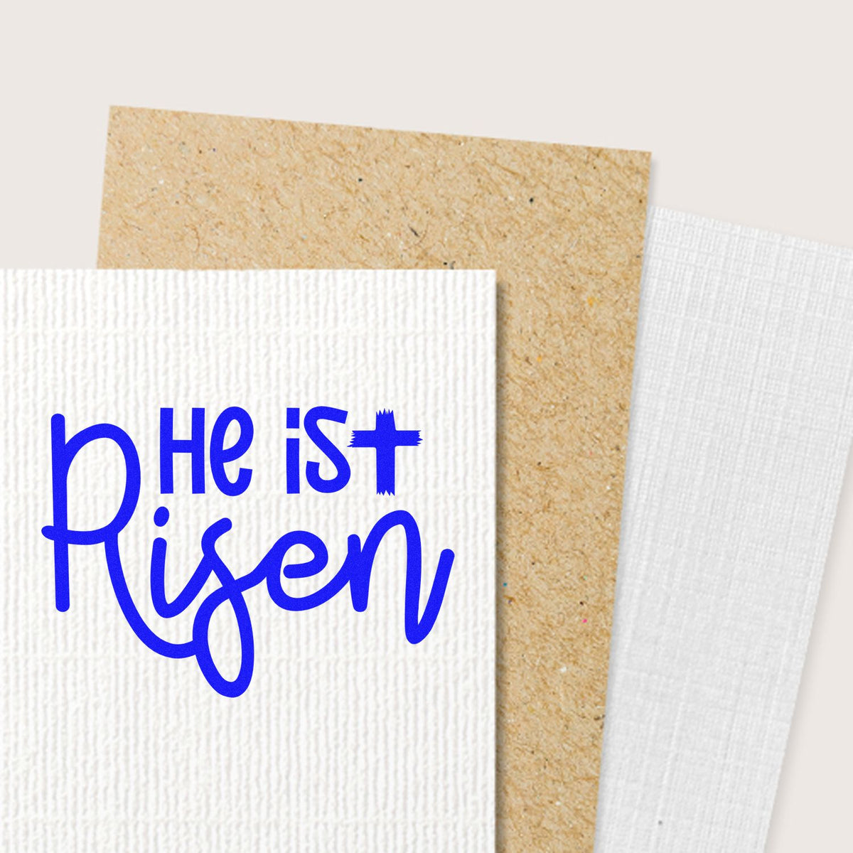 Blue He Is Risen stamp on white textured cardstock, shown with tan kraft and white seal paper samples.