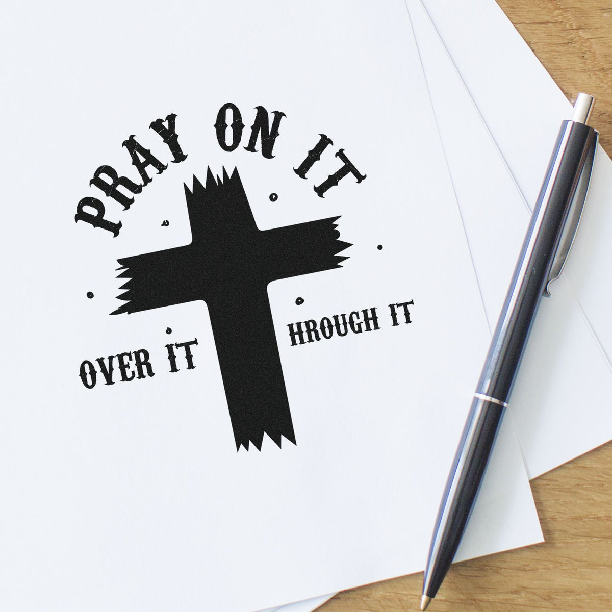 Black faith-based stamp and seal design with cross and Pray On It, Over It, Through It on white paper, beside a silver pen.