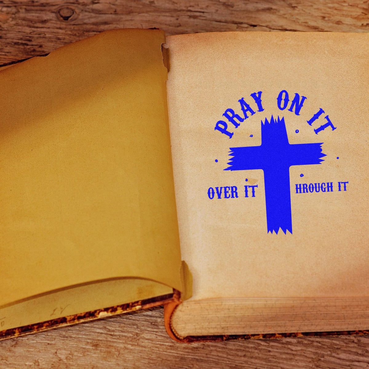 Open tan book with a blue cross stamp reading Pray on it, over it, through it on rustic wood.