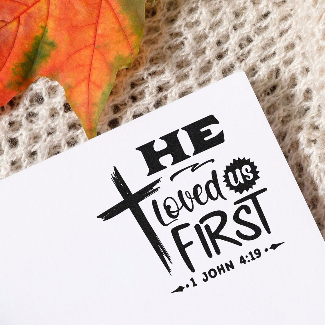Black stamp and seal design reading He loved us first with a cross on white paper, beside an orange autumn leaf.