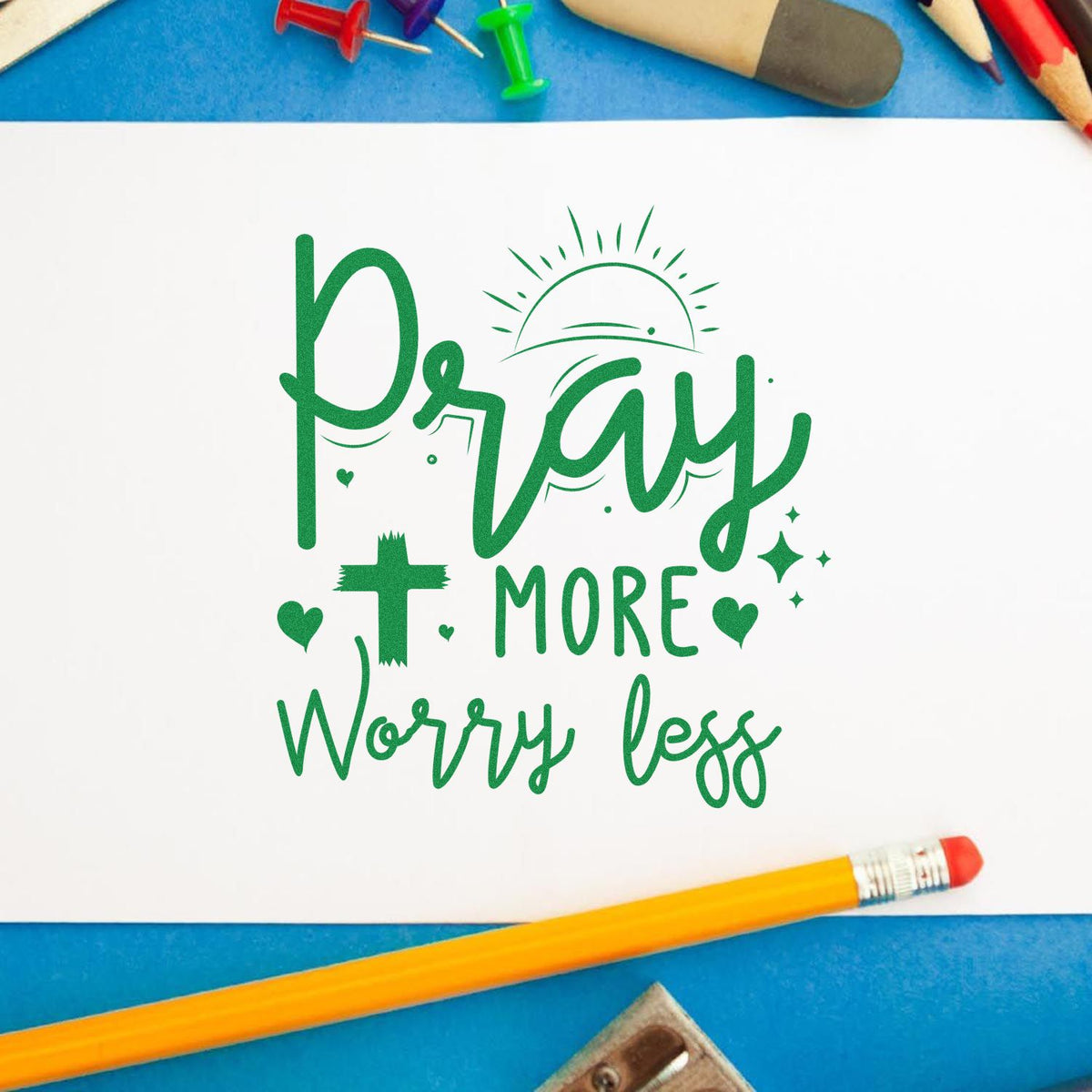 Green Pray more Worry less stamp-and-seal design on white paper, with pencils and push pins on a blue background.