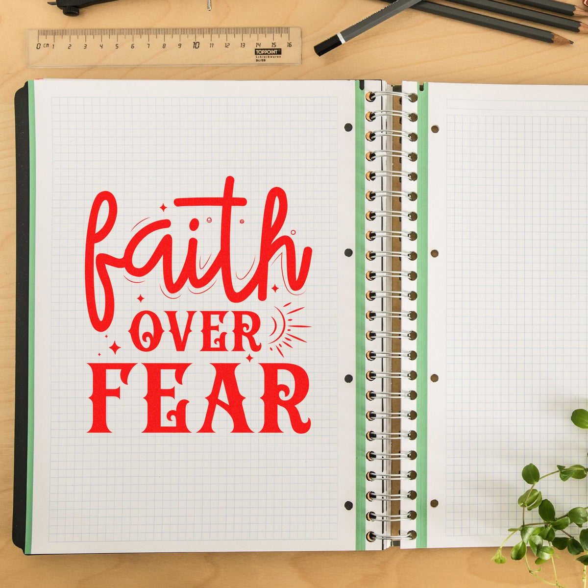 Red faith over fear rubber stamp seal on a grid notebook page, with spiral binding, ruler, pencils, and green leaves.