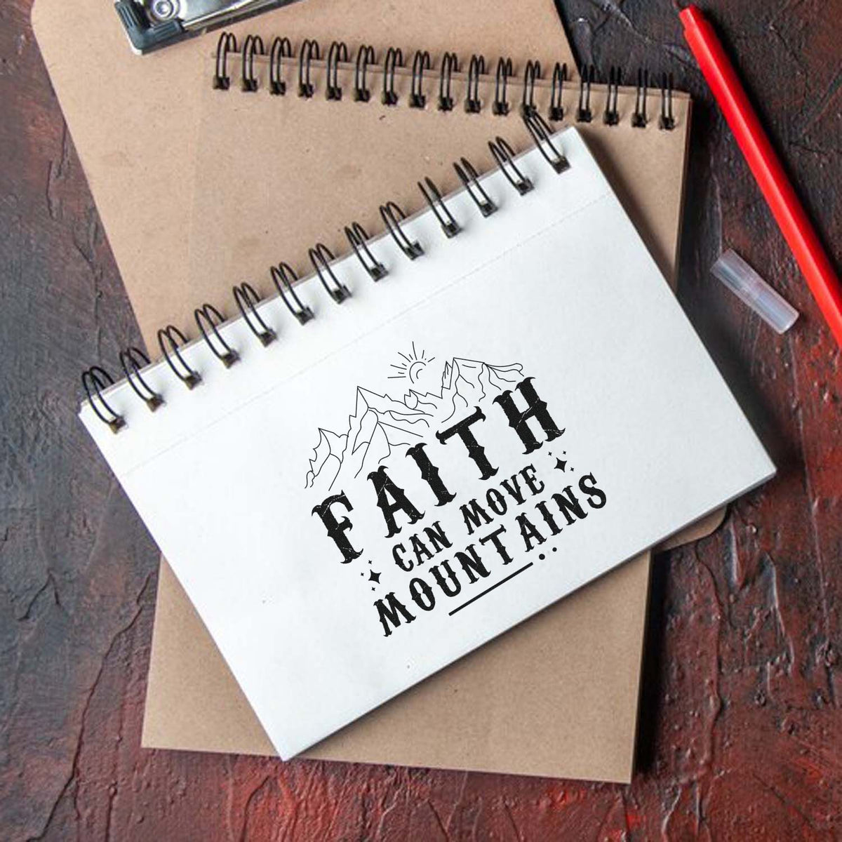 White spiral notebook with black Faith Can Move Mountains stamp/seal design, layered over brown kraft notebook, beside a red pen.