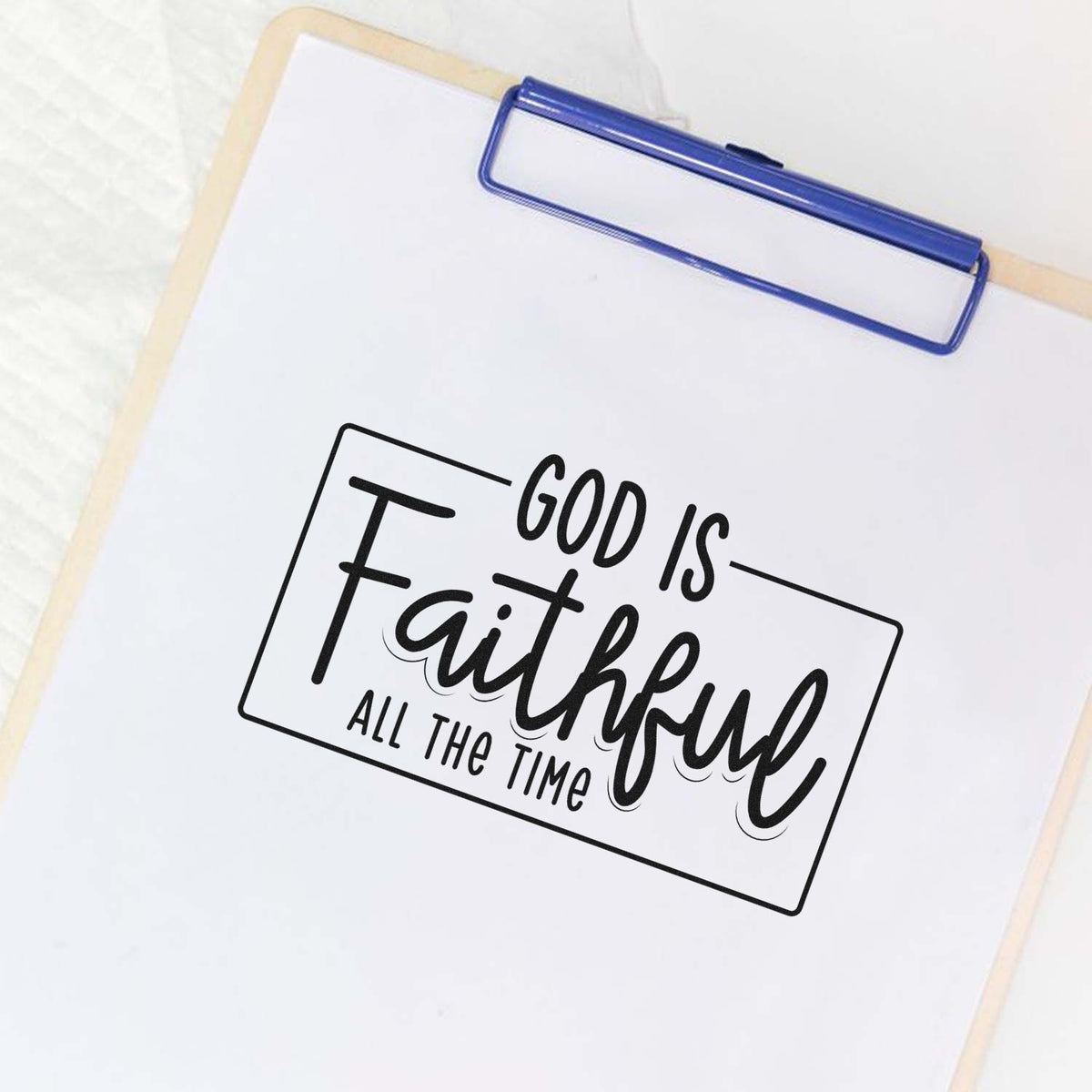 Blue clipboard holding a black faith stamp/seal design reading God Is Faithful All the Time on white paper.