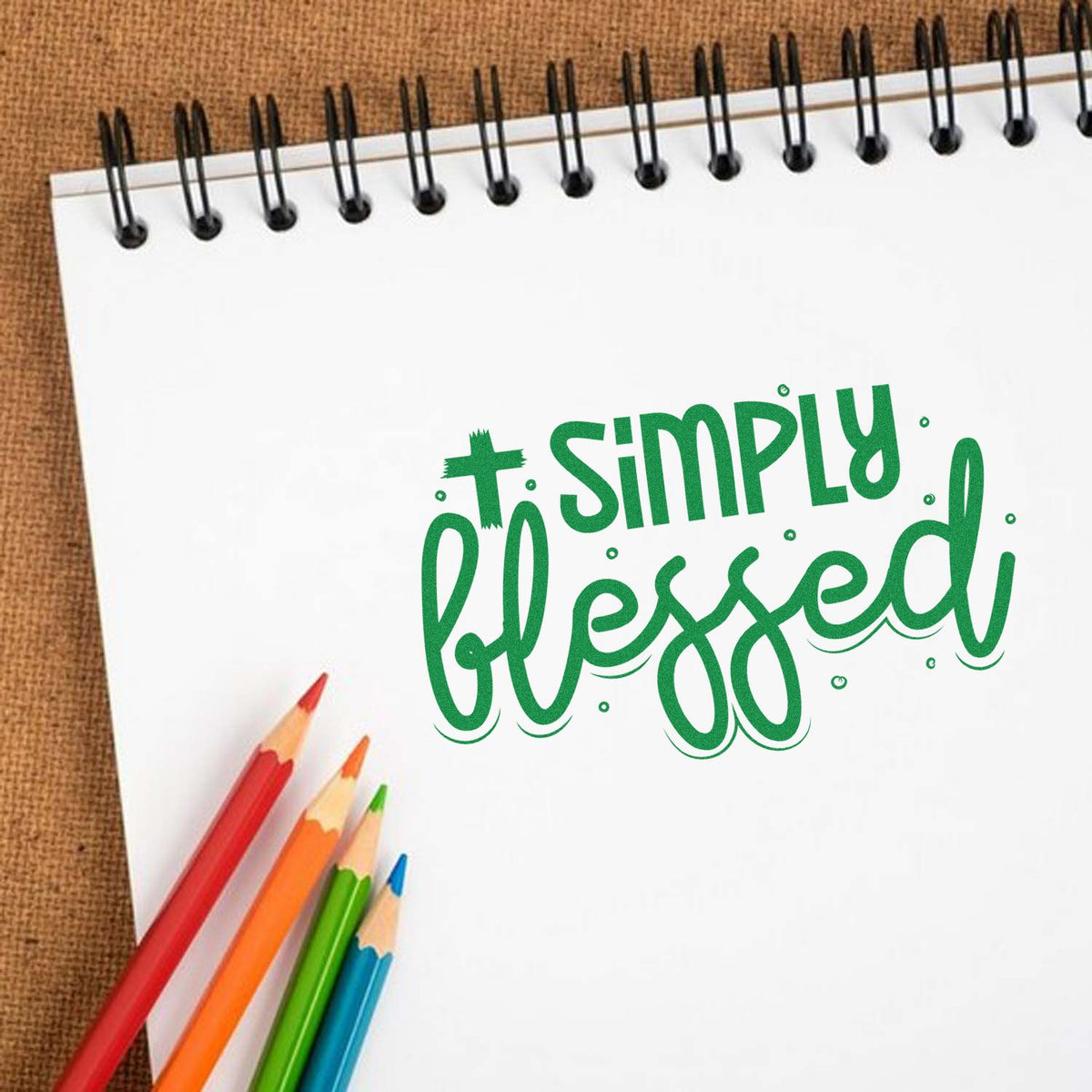 Green Simply blessed rubber stamp design on a spiral notebook with colorful pencils on a brown fabric background