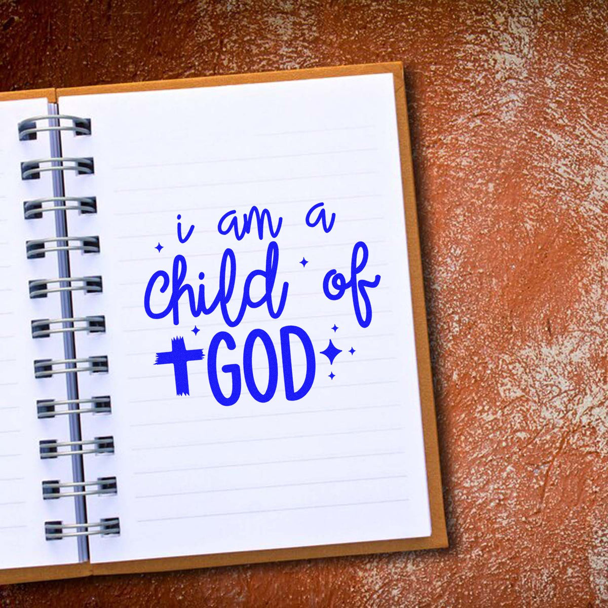 Blue faith-themed stamp seal reading i am a child of GOD on a white spiral notebook page with a brown textured background.
