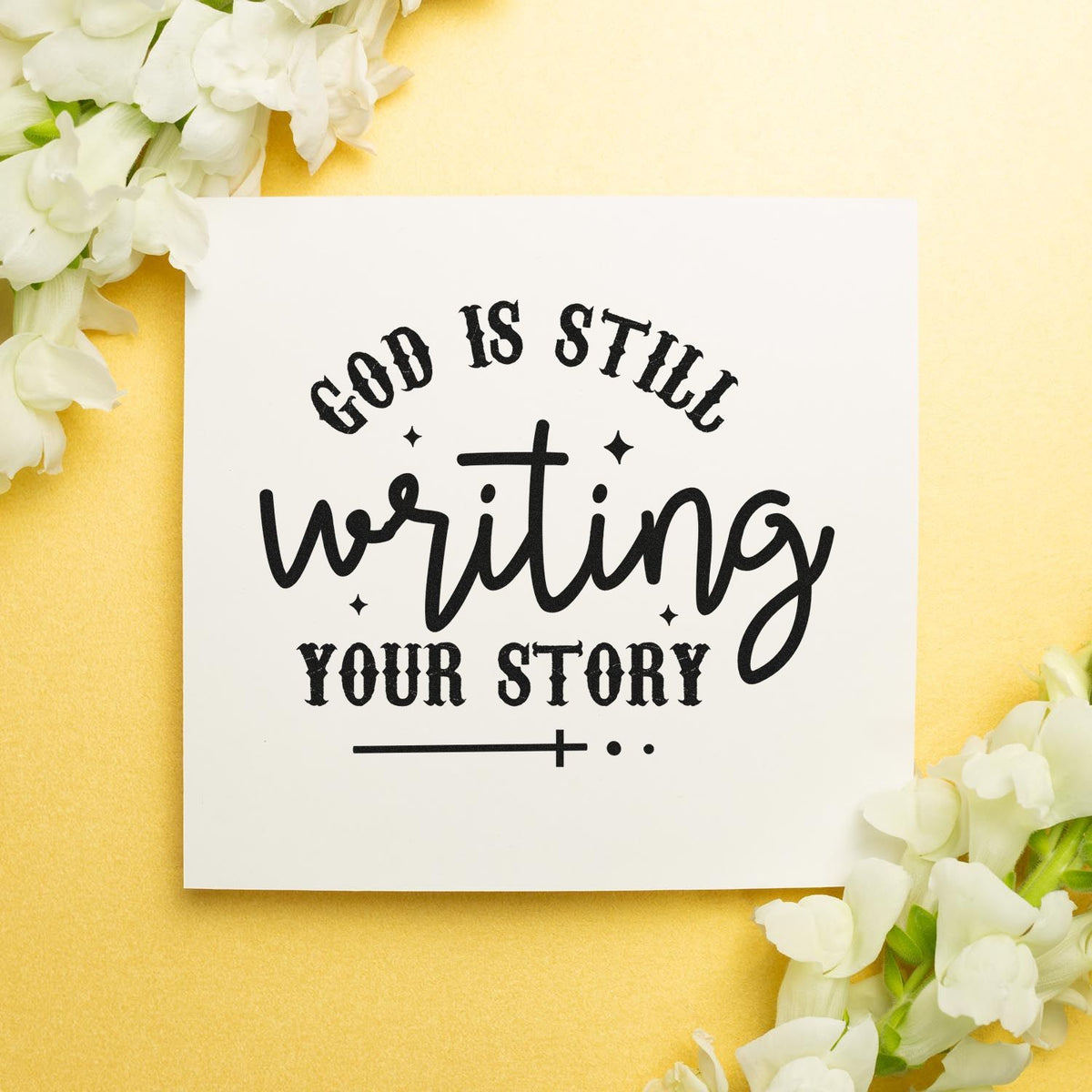 Black stamp seal reading God is still writing your story on a white card with white flowers and warm yellow background.