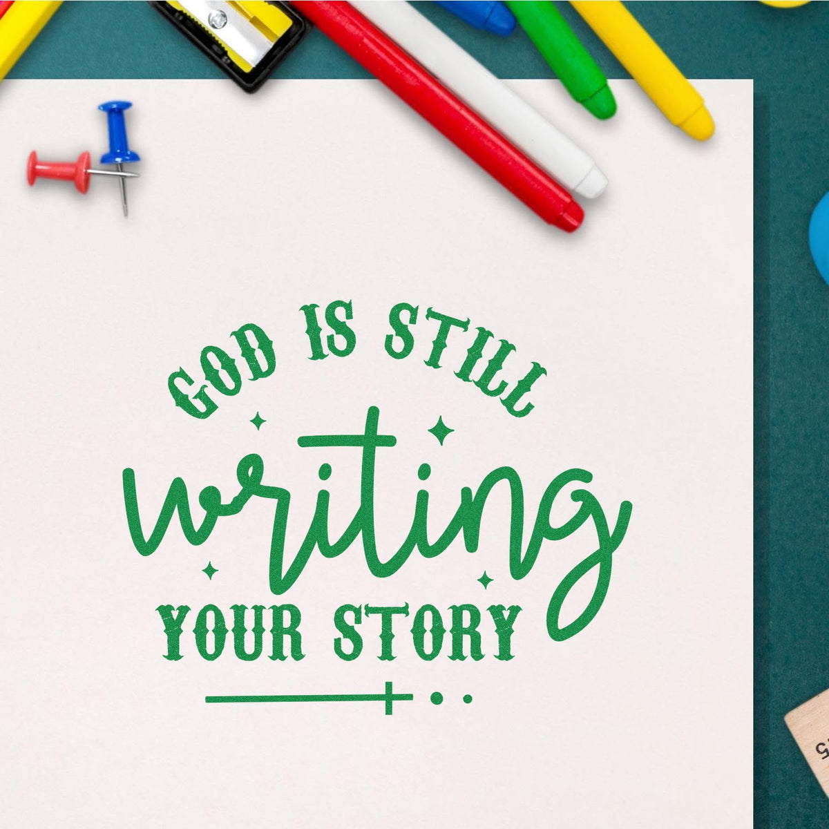 Green rubber stamp and seal reading God is still writing your story on white paper, with colorful pens and push pins around it.