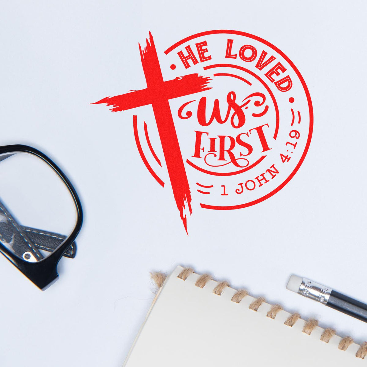 Red Christian rubber stamp seal with cross and He Loved Us First, 1 John 4:19 on white paper.