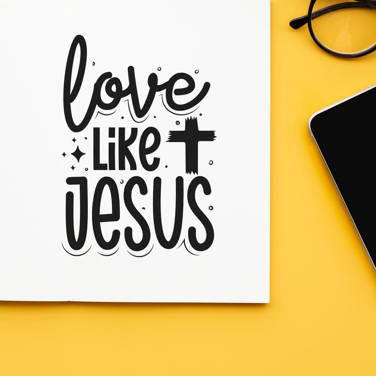 Black decorative stamp-style Love Like Jesus seal on white paper, set against a bright yellow background with black glasses.