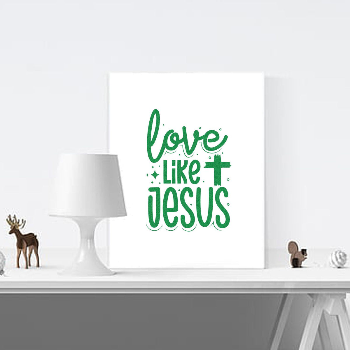 White wall decor with green stamp-style Love Like Jesus lettering, shown beside a lamp and small shelf decor.