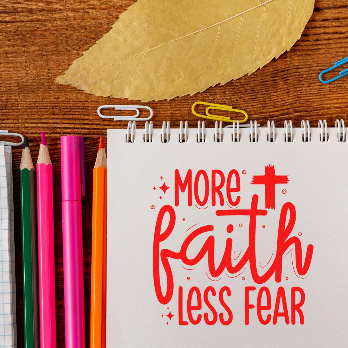 Red rubber-stamp-style More Faith Less Fear on a white notebook, with colorful pencils, paper clips, and a leaf on wood.