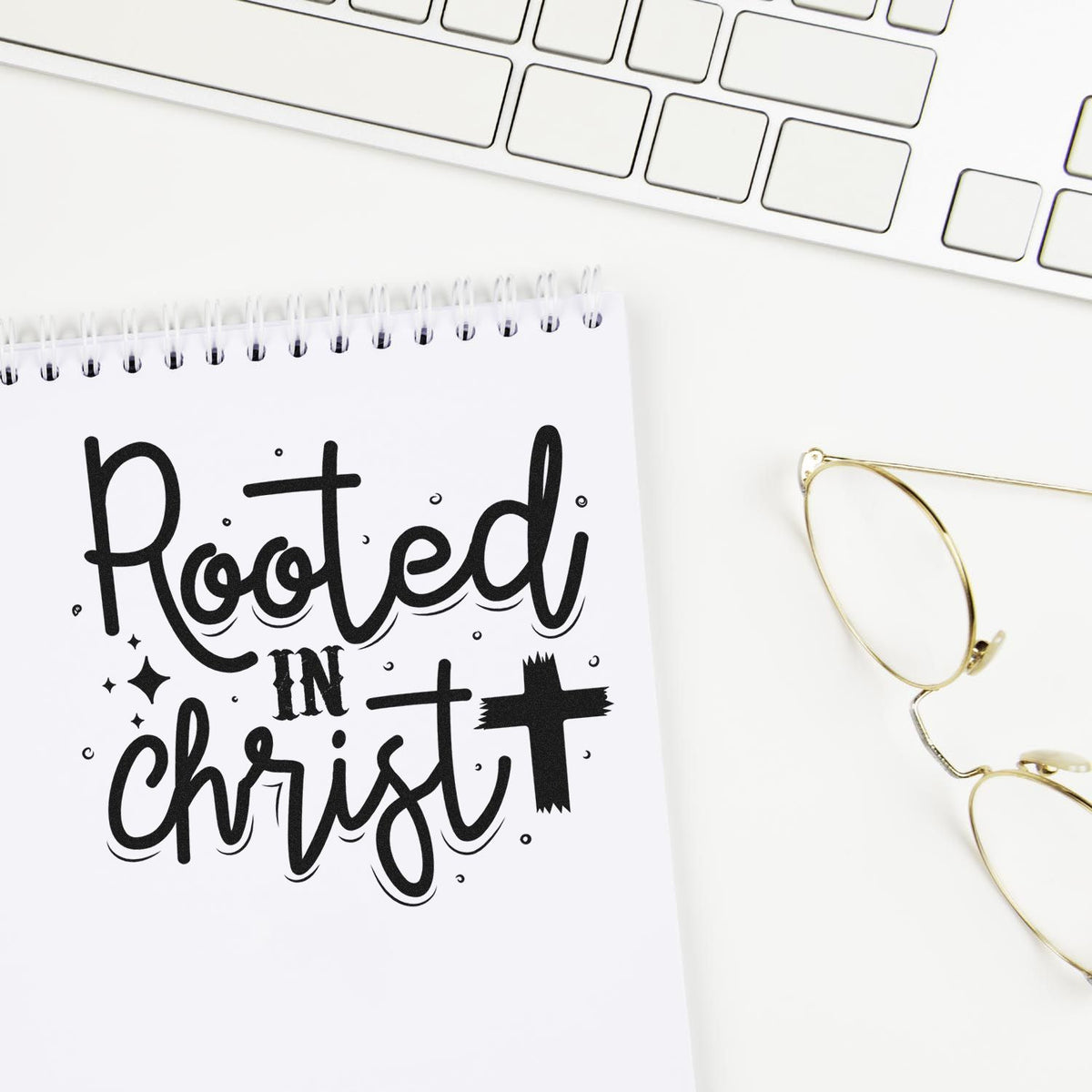 Black Rooted in Christ stamp and seal design on a white spiral notebook beside a keyboard and gold wire-frame glasses.