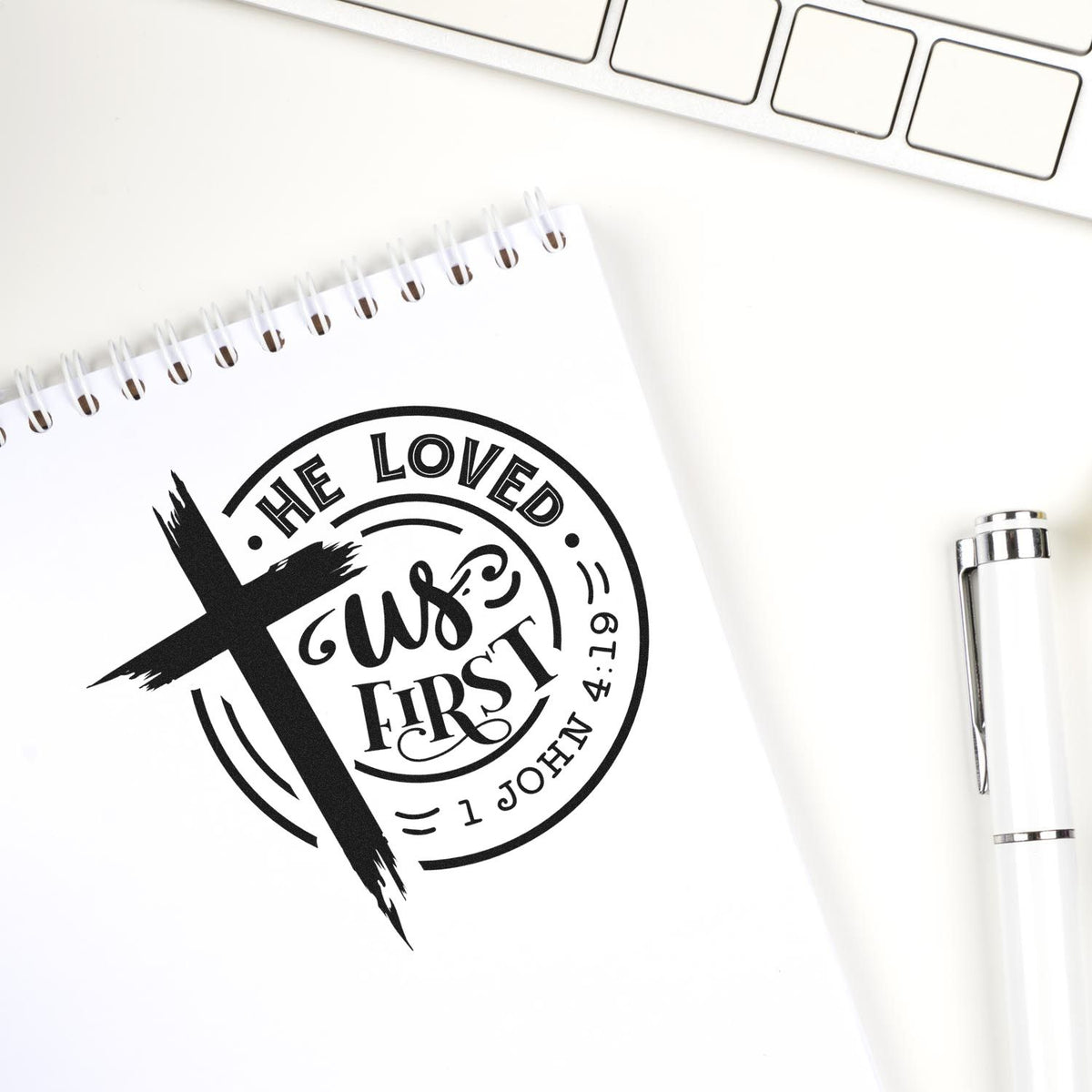 Black custom stamp seal with cross and He Loved Us First on a white notepad, beside a white pen and keyboard.